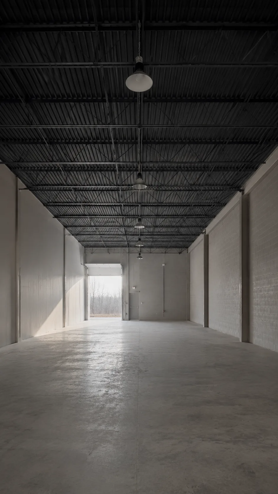 An empty light-industrial unit, clear span, high-bay fixtures overhead and the loading door open at the far end.